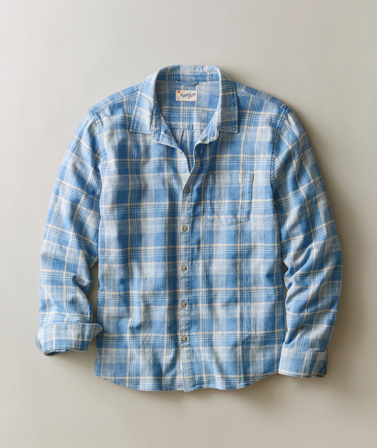 A blue and white plaid long-sleeve button-up shirt laid flat against a neutral light gray background.
