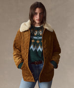 A woman wearing a brown quilted jacket with a cream sherpa collar, layered over a green and white patterned knit sweater, blue jeans, and a brown belt, set against a neutral textured studio background.